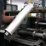 Bediener handhabt ein zylindrisches Metallbauteil während des Rollformprozesses auf einer industriellen Rollformmaschine.